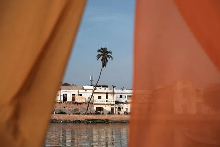 透过前景两侧橙色布料形成的狭长缝隙取景，中景可见一棵微微倾斜的棕榈树立在河岸边，后方是一排色调斑驳的低矮建筑，外墙带有岁月痕迹与不规则修补痕迹。前景布料略微失焦，形成自然的框景效果，引导视线集中在棕榈树与河岸建筑上。下方河面映出建筑与天空的倒影，水面平静，整体画面呈现安静而温暖的日常风景，强调在缝隙中被捕捉到的光与空间层次。林道锦《谁在缝隙里种光》