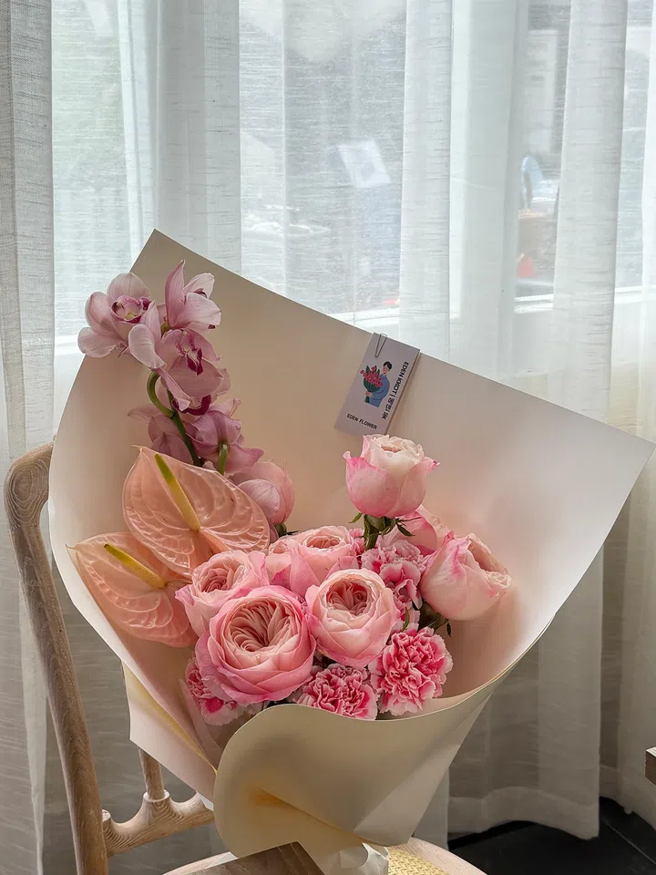 一束粉色系花束摆放在木质椅子上，背景为透光白色窗帘与自然光线，整体画面呈现清新而优雅的氛围。花束以多种粉色花卉组成，包括盛开的粉色玫瑰、兰花、康乃馨与粉色火鹤花，花瓣层次丰富，色调从浅粉到柔和玫瑰粉渐层变化，展现细腻质感。花束外层以浅米色包装纸包裹，并附有 Eden Flower 品牌卡片，增添精致送礼感。自然光从窗边洒入，使花瓣纹理更加清晰，强化整体柔和色调与浪漫气息。整张图片突显花束作为经典情人节礼物的优雅与表达心意的象征。2026情人节礼物推荐：10款精致礼物让TA一秒心动