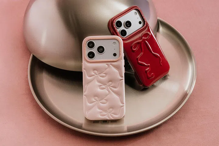 两款 CASETiFY Pillow Case 手机壳摆放在金属圆盘上，背景为粉色桌面，整体呈现精致且富有情人节氛围的视觉效果。手机壳分别为柔和的粉色与亮面红色，两者均采用立体蝴蝶结设计，蝴蝶结纹理微微隆起，呈现出仿枕头般的蓬松质感，增添可爱与时尚感。粉色款手机壳为哑光柔和质地，散发优雅气息；红色款则为亮面设计，在光线下呈现出光泽感，更显醒目。两款手机壳均安装在手机上，可见背部镜头开孔设计完整贴合机身。整体画面以粉色与金属色调为主，营造出甜美又现代的情人节礼物氛围，突显手机壳作为兼具实用性与设计感的送礼选择。2026情人节礼物推荐：10款精致礼物让TA一秒心动