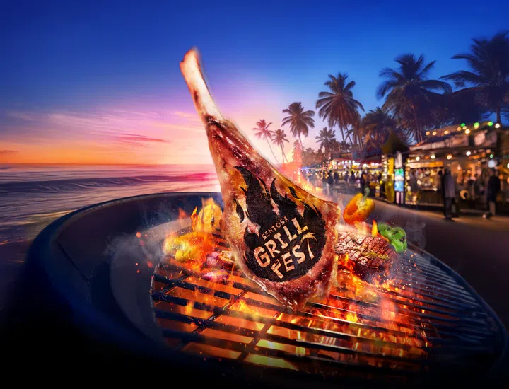 一块印有“Sentosa GrillFest”标志的巨大带骨牛排正在炭火烤架上炙烤，火焰四起，周围摆放着彩椒、玉米及牛排等烧烤食材，营造热腾腾的烧烤氛围。背景是Siloso Beach的海边夕阳景色，椰树林立，远处可见热闹的美食摊位和来往人群，在橙紫色晚霞映衬下，展现Sentosa GrillFest结合海滩、美食与夏日夜晚的热闹氛围。