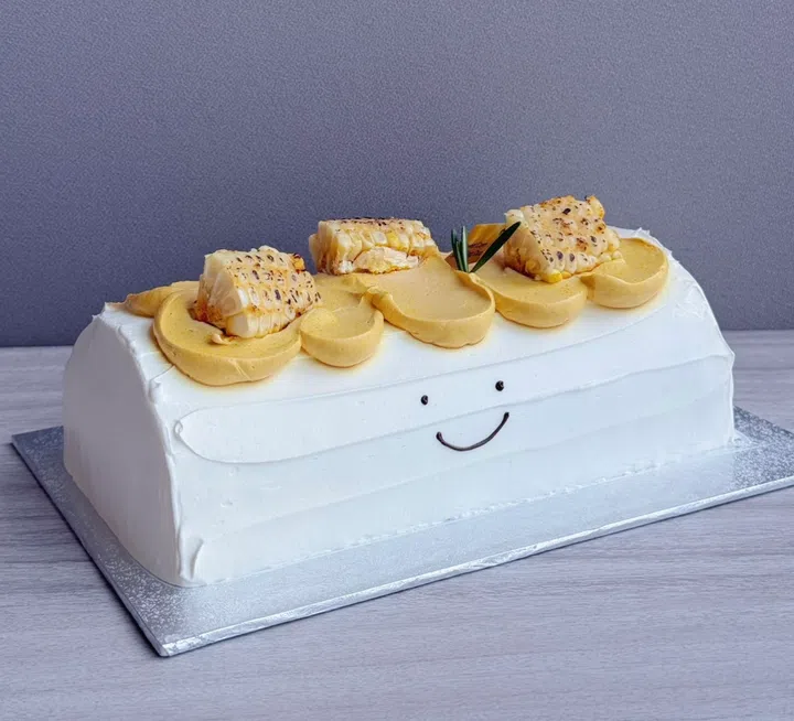 The Pine Garden 的 Sweet Corn Salted Egg Duo Log 圣诞蛋糕，以白色鲜奶油外层搭配玉米与咸蛋黄奶油装饰，并点缀烤玉米块，风味呈现香甜玉米与咸蛋黄的平衡层次，蛋糕正面带有可爱笑脸设计。2025 新加坡圣诞蛋糕推荐｜好吃又好看的 Christmas Cakes & Logcakes 精选清单