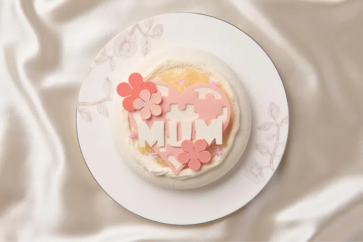 白色圆盘上摆放一款简约精致的蛋糕，表面覆盖细腻浅色慕斯，中央装饰粉色心形巧克力与花朵元素，并拼出“MUM”字样，整体色调柔和，以奶油白与樱花粉为主，搭配丝绸质感背景，呈现温馨优雅的母亲节氛围。2026母亲节蛋糕推荐：精致又好吃的限定款