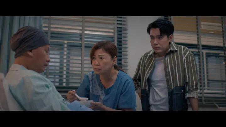 《你好，再见》剧照。向云饰演的徐巧妹在病房内细心照顾患癌丈夫，手中拿着碗匙耐心喂食，神情担忧而疲惫，儿子则站在一旁默默陪伴，画面充满家庭面对病痛时的沉重氛围。