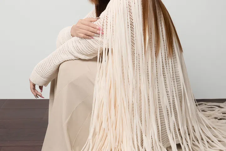 Yumiko Kayahara 坐在木质地板上，身穿米白色长流苏针织外套与浅色长裙，神情平静而专注，背景简洁明亮，展现柔和而自信的气质。