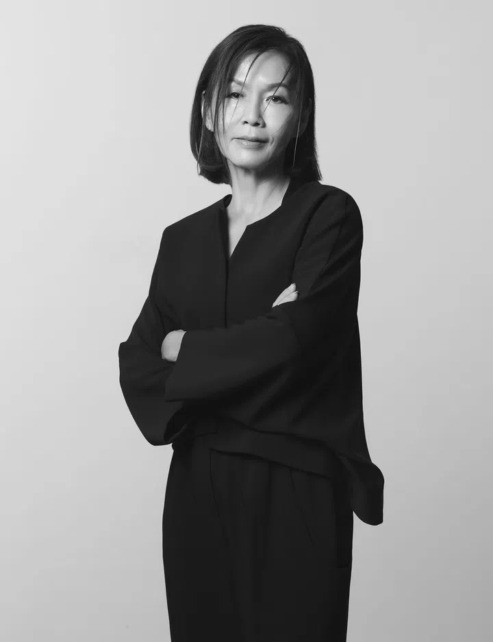 一张黑白人物肖像照，一位女性站在浅灰色背景前，以正面姿态面对镜头。她留着及肩长度的短发，几缕发丝自然垂落在脸侧，增添轻微随性感。她双手交叉抱在胸前，神情平静而自信，嘴角带着淡淡笑意，眼神柔和地看向镜头。身穿一套简约深色服装，上衣为宽松剪裁设计，袖口较宽，搭配同色长裤，整体线条流畅俐落。黑白色调让画面更显沉稳与层次感，也突显人物成熟、从容而坚定的气质。