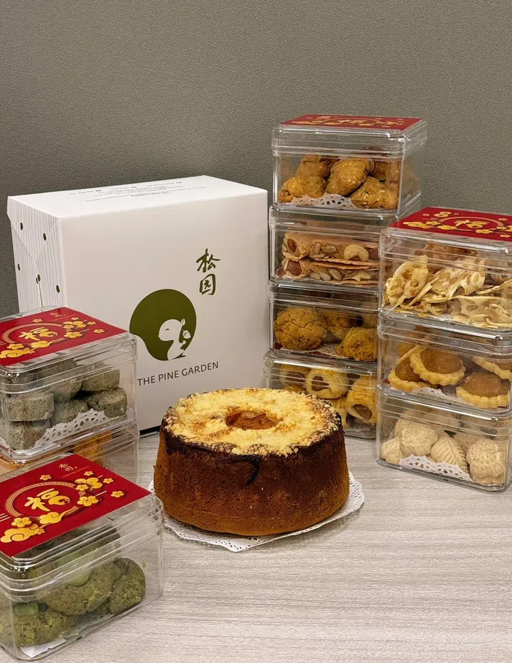 画面展示 The Pine Garden 新春年饼与节庆蛋糕组合陈列在浅色木质桌面上，整体呈现传统与现代并存的年节送礼场景。中央是一款圆形戚风蛋糕，顶部微微开裂并露出浅黄色蛋糕组织，放置在白色蕾丝纸垫上，突出蛋糕松软蓬松的质地。后方摆放印有 The Pine Garden 标志与中文“松园”字样的白色礼盒包装。四周整齐堆叠多盒透明方形年饼盒，内装不同种类的新年点心，包括凤梨酥、曲奇饼、坚果饼干、爱心酥与传统白色年饼等，盒盖贴有红色新春贴纸，增添节庆感。左前方另有装着绿色口味饼干的年饼盒，整体呈现适合拜年与家庭聚会分享的多款年饼与蛋糕选择。文章标题：“2026马年年饼推荐：新春送礼与拜年点心精选”。