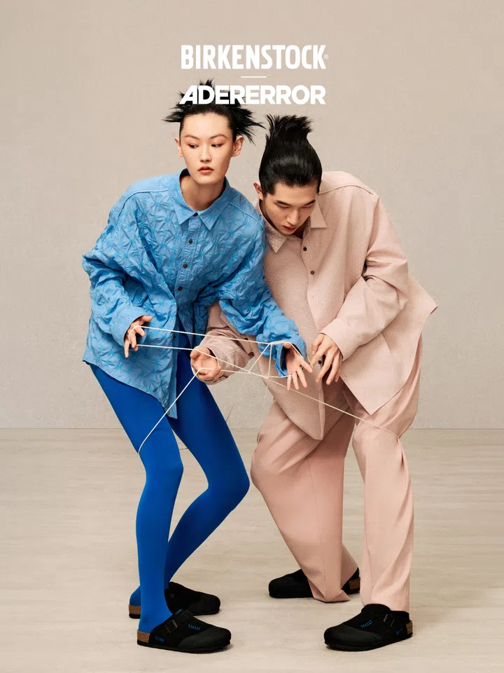BIRKENSTOCK x ADERERROR联名宣传照，两名模特站在米色背景前，以白色细线缠绕双手，呼应此次联名以“Thread”为主题的设计概念。左侧模特身穿亮蓝色宽松衬衫和同色紧身裤，右侧模特则穿着浅粉色宽松西装造型，两人脚上都搭配黑色BIRKENSTOCK Boston联名鞋款，鞋面可见蓝色缝线细节。画面上方印有BIRKENSTOCK与ADERERROR品牌标志，整体通过蓝、粉与黑色的鲜明对比，突出联名系列兼具实验感与时尚感的视觉风格。