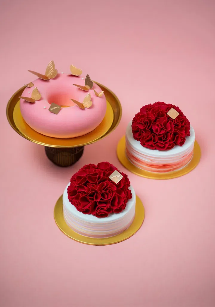 粉色背景上摆放三款精致蛋糕，包括一款粉色环形慕斯蛋糕，表面点缀金色蝴蝶装饰，以及两款顶部覆盖立体红色花瓣造型的圆形蛋糕，整体配色柔和浪漫。2026母亲节蛋糕推荐：精致又好吃的限定款