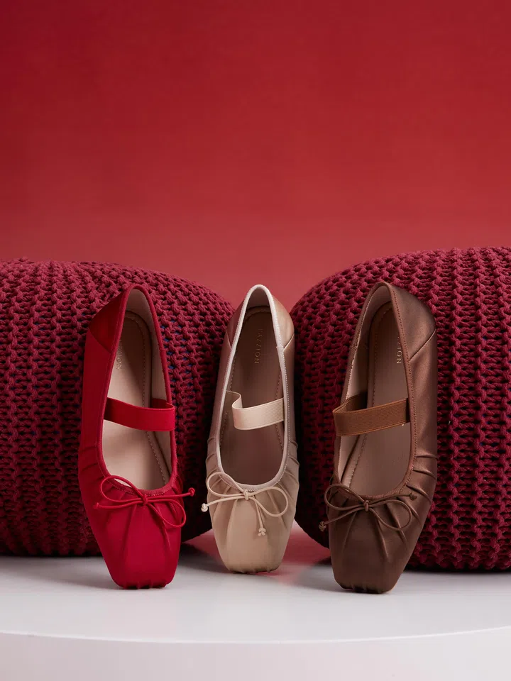 图片展示三双不同颜色的PAZZION Brivelle Silk Mary Janes平底鞋，整齐摆放在白色圆形台面上。三双鞋分别为鲜艳红色、柔和米色与沉稳棕色，鞋款均采用经典Mary Jane设计，配有弹性鞋带与精致蝴蝶结细节，展现优雅甜美的风格。鞋面材质呈现丝质光泽质感，线条柔和流畅，方头轮廓设计让整体外型更显时尚。背景为温暖的红色渐层色调，两侧摆放红色针织抱枕作为装饰，营造浓厚的新年节庆氛围。画面整体风格简约精致，突显鞋款的多色选择与舒适优雅的设计特点，非常适合新年拜年与日常穿搭；2026新年鞋推荐：舒适好穿又好看的拜年鞋款。