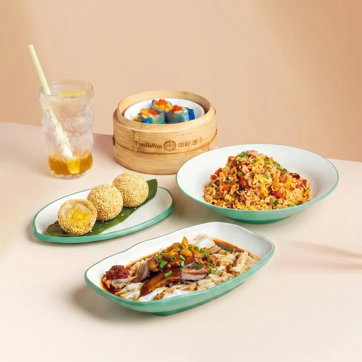 浅色背景前摆放多道 Tim Ho Wan 国庆限定港点，包括肉脞肠粉、培根午餐肉 XO 炒饭、芋泥金沙煎堆，以及蓝色外皮的叻沙海鲜饺，旁边搭配一杯清爽饮品。整体以简洁明亮的风格呈现，将新加坡本地风味与港式点心结合。
