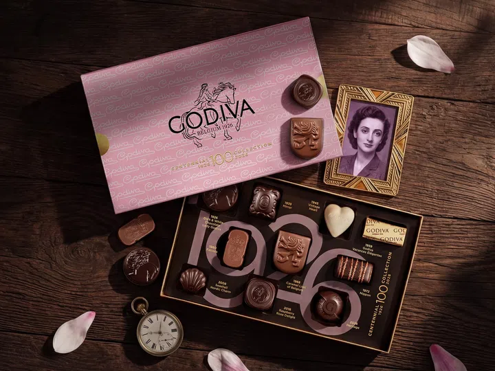 GODIVA Centennial Chocolate Gift Box 百年纪念巧克力礼盒产品照，以复古木质桌面搭配粉色礼盒包装呈现浓厚复古与优雅氛围。画面中展示多款经典 pralines 巧克力，包括心形、贝壳与雕花设计巧克力，并加入怀表、旧照片与花瓣元素，呼应品牌100周年历史与比利时巧克力工艺。整体风格精致浪漫，非常适合作为母亲节送礼、下午茶甜点或收藏系巧克力礼盒推荐。母亲节礼物推荐2026：从实用家居到时尚科技，这些礼物妈妈真的超爱！