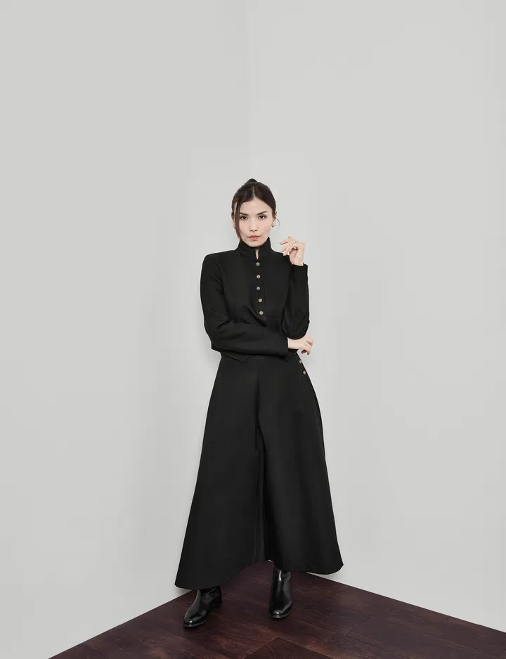 Rachel Mak 为《The Millennial Talk》专访拍摄的肖像照，她身穿黑色高领长袖连身裤，搭配黑色短靴，双臂自然交叠站立在深色木质地板上，一手轻触脸侧，目光直视镜头。背景为简洁的浅色墙面，整体构图干净对称，呈现她从模特身份走向创办色彩分析品牌 Niji Colours 的沉稳与专注状态。