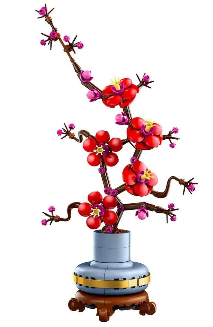 画面展示 LEGO Icons 傲骨梅（Plum Blossom，10369）积木盆栽模型。主体为一株梅花造型植物，由深棕色枝干积木构成弯曲伸展的树枝结构，枝头盛开多朵红色梅花，并点缀紫粉色花蕾与黄色花芯细节，呈现层次丰富的开花状态。枝条间还可见尚未完全绽放的花苞，增强自然生长感。下方为圆形蓝灰色花盆，外圈搭配金色装饰环，并置于深棕色底座上，使整体造型稳固且具有东方花艺风格。背景为纯白底，突出植物主体的色彩与线条轮廓。这款积木盆栽适合作为农历新年或日常居家空间的装饰摆设，也象征坚韧与新生的寓意。