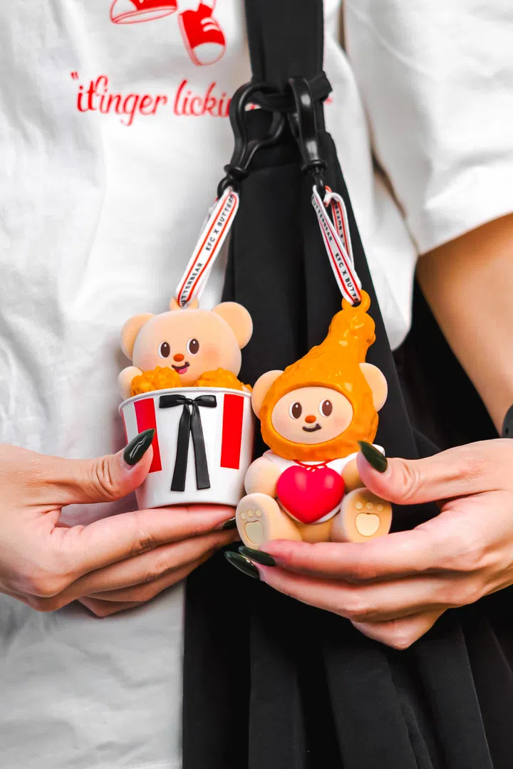 特写画面中，一位手持两只KFC × Butterbear联名公仔的人物，左边是抱着炸鸡桶的熊，右边是戴着橙色“辣酱”造型头套、抱着红心的熊，造型可爱讨喜。