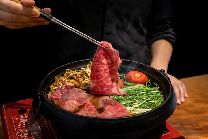 Fat Cow母亲节限定寿喜烧套餐图：食客正以筷子夹起油花细致的和牛片放入热腾腾的寿喜烧锅中，锅内可见时令菇类、水菜与番茄等食材，整体画面充满浓郁和牛香气与温暖日式料理氛围，展现精致又带点奢华感的母亲节用餐体验。2026母亲节餐厅精选指南
