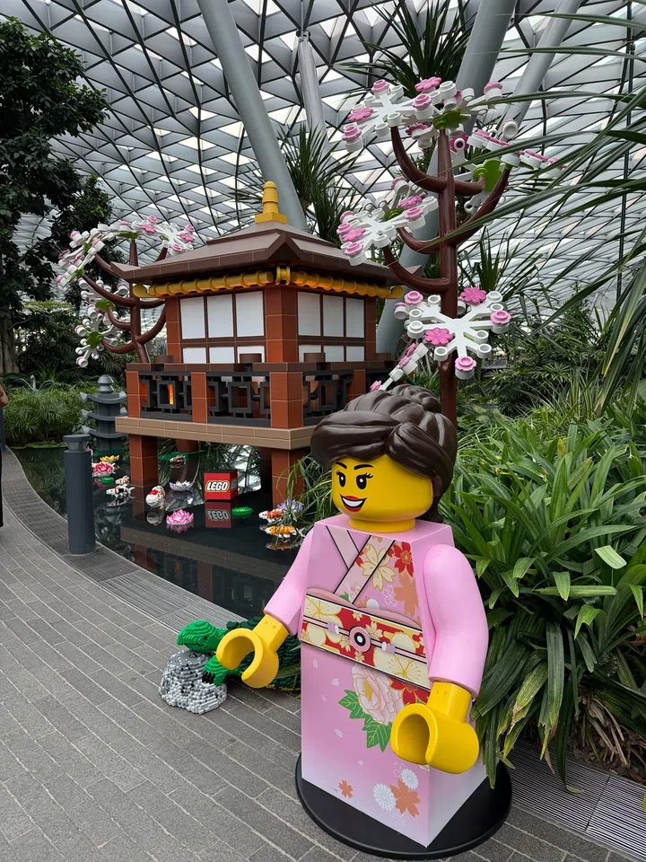 Jewel Changi Airport的Forest Valley展示一座以LEGO积木打造的日式庭园场景，包括一座日式凉亭、盛开的樱花树及水面上的LEGO荷花装置。前方摆放一尊身穿粉色和服的LEGO人偶雕像，四周环绕绿植、水景与玻璃穹顶，呈现“Celebrate Jewel Blooms with LEGO Botanicals”活动中的日式花园主题展示。
