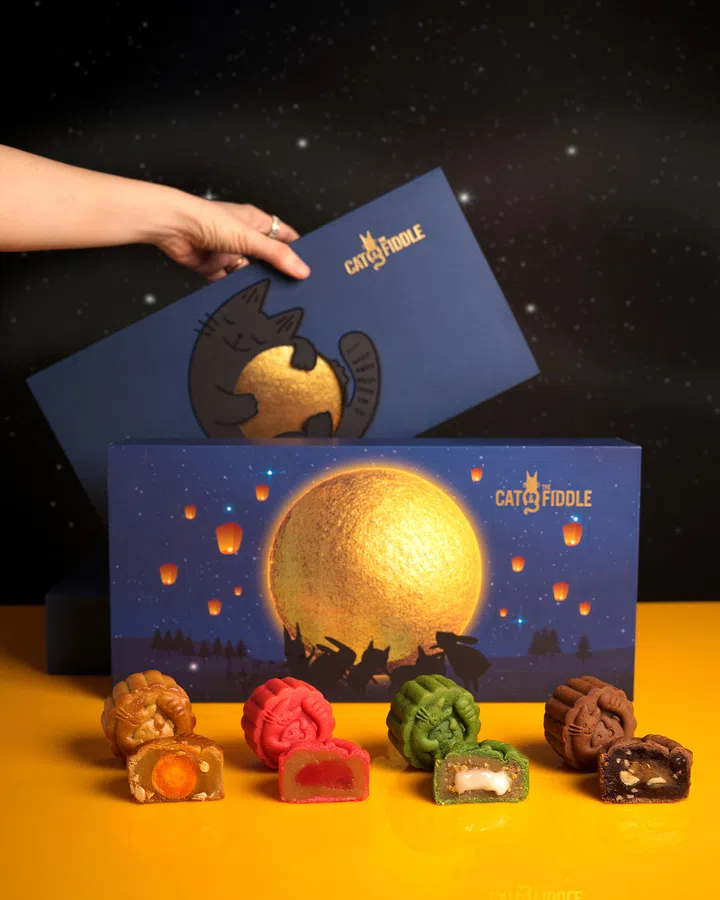深蓝色的 Cat & the Fiddle 月饼礼盒以猫咪抱月与金色圆月为主视觉，点缀夜空与天灯图案。前方摆放四枚不同口味的烘焙月饼，分别为金黄、红色、绿色与棕色，其中两枚对半切开，露出咸蛋黄、莲蓉、果馅与坚果内馅。背景为星空设计，整体氛围俏皮又温馨。Cat & the Fiddle，2025中秋月饼必买推荐。