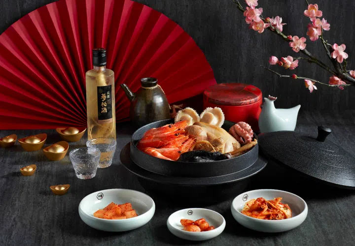 一锅丰盛的韩式滋补海神汤置于黑色石锅中，锅内盛有整只鸡、鲜橙色大虾、鲍鱼与章鱼等海陆食材，汤料丰富饱满。石锅旁摆放韩国人参酒与小酒杯，搭配传统陶壶与白色小碟装泡菜及配菜。背景以红色折扇、粉色梅花枝与红色圆盒点缀，营造浓厚农历新年节庆氛围，整体呈现适合新春期间与家人共享的滋补佳肴与聚餐场景。新加坡大年初一初二照常营业餐厅推荐：8家不打烊好去处（2026马年）