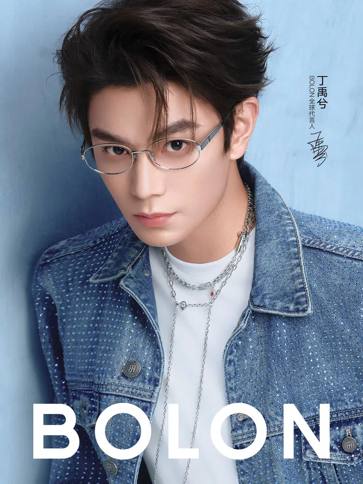 男艺人身穿蓝色牛仔外套搭配白色圆领上衣，配戴多层银色项链，佩戴银色金属椭圆形镜框，直视镜头。发型蓬松有层次，整体造型干净利落。镜框采用细金属设计，线条简约轻盈，突显五官轮廓。画面背景为蓝色渐层，并印有BOLON品牌标志及人物签名字样。