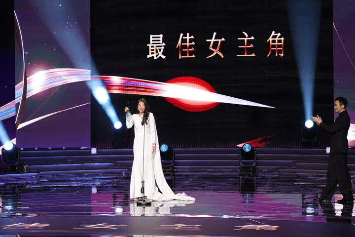 这是一张在《红星大奖》颁奖典礼现场拍摄的全景画面。舞台中央，刘子绚身穿一袭白色长礼服，裙摆与拖尾自然铺展在地面上，站在麦克风前，手中举起绿色奖座，脸上带着喜悦而从容的笑容，成为全场焦点。
舞台背景以黑色为主，上方清晰显示“最佳女主角”字样，搭配红色与白色的光影线条横跨画面，形成强烈的视觉冲击。多束聚光灯从舞台两侧打下，照亮中央位置，同时在光滑的地面上形成清晰的倒影，增强整体空间感与仪式感。
画面右侧，一位身穿黑色西装的男艺人正面向刘子绚鼓掌致意，神情带着欣赏与祝贺，进一步烘托出她获奖时刻的荣耀与肯定。舞台台阶与灯光设备清晰可见，整体构图开阔而有层次，完整呈现出她在颁奖典礼上获奖的高光瞬间。刘子绚首夺视后：22年7次提名终圆梦，她把时间走成答案