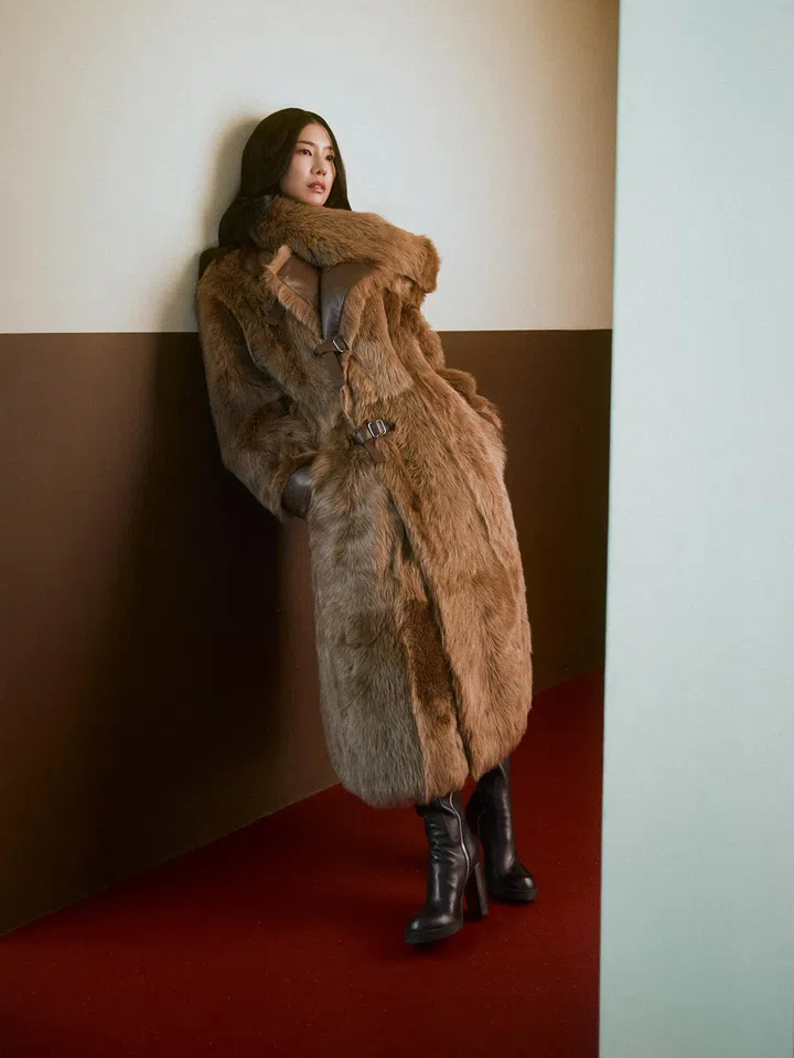 Ferlyn身穿棕色长款毛绒大衣与黑色长靴，倚靠在室内墙边，神情平静，整体画面简洁而带有冷感氛围。