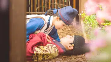 一名身穿蓝色传统韩服的女性俯身靠近躺在地上的男性，两人四目相对，眼神温柔而专注。男性身穿红色与金色刺绣的韩服，神情平静而深情。画面背景为古代庭院一角，周围有木质围栏与落叶，前景点缀柔和粉色花朵，整体氛围浪漫唯美，呈现出韩剧中经典的古装爱情场景与强烈的 CP 感。暴君的厨师|2025韩剧最强CP是谁？看完只想喊——原地结婚吧！