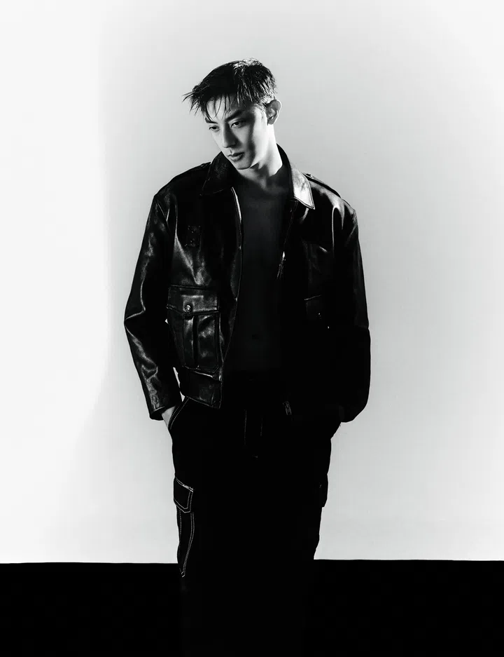 仲伟杰黑白全身人像，身穿皮革外套与深色长裤，微微低头站立于简洁背景前，整体风格随性且富有时尚感。