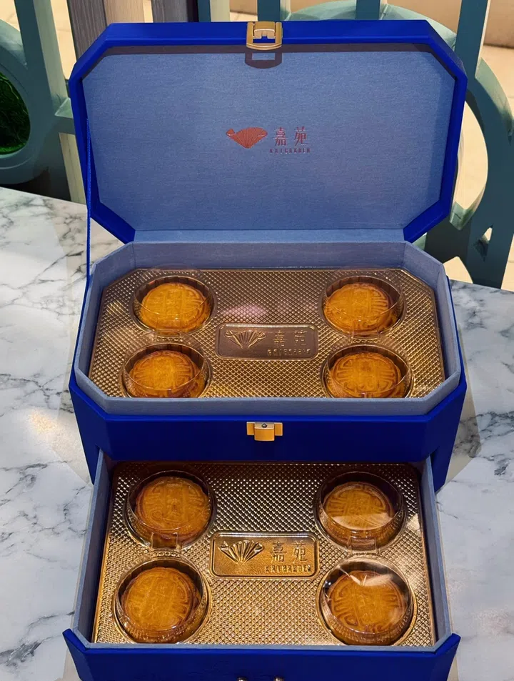 KAI GARDEN（嘉苑）双层宝蓝色礼盒置于大理石桌面，礼盒内部金色衬底上整齐摆放六枚金黄色迷你奶黄流沙月饼。礼盒设计奢华大气，中央刻有嘉苑标志与扇形图案，整体呈现典雅高贵的格调。KAI GARDEN（嘉苑），2025中秋月饼必买推荐。