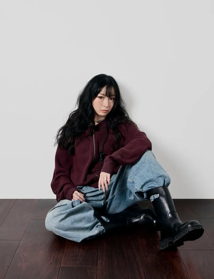一位女性坐在深色木地板上，背靠简洁的浅灰色背景，整体画面干净利落。她留着黑色长卷发与齐刘海，妆容精致，眼神沉稳中带点冷静的个性，直视镜头。她身穿酒红色连帽卫衣，搭配宽松浅蓝色牛仔裤与黑色皮革中筒靴，造型偏街头风又带一点随性态度。姿态自然，一腿弯曲、一腿伸展，手轻放在膝上，整体呈现出自在却有力量感的氛围，展现出独立、自我且不受拘束的个人风格。