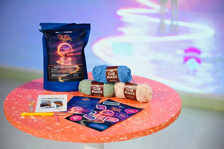 Geneco与The Club Made合作推出的限量版“Begin Again”钩织入门礼包（Crochet Starter Kit）陈列于圆形展示桌上。礼包内包含钩针、三卷不同颜色的毛线、编织教学说明、小配件及主题贴纸等材料，适合初学者体验钩织创作。深蓝色包装袋印有“Begin Again”主题设计，背景可见展览现场的灯光装置与柔和光影，呼应企划鼓励大众透过钩织放慢脚步，在创作过程中重新认识自己、迎接新的开始。