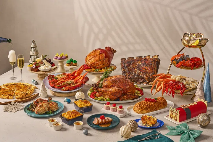 一整桌 JW Marriott Singapore South Beach 的 Beach Road Kitchen 节日自助餐料理，包括龙虾、螃蟹、海鲜塔、烤火鸡、枫糖烤火腿、烤肋排、披萨、配菜与多款圣诞蛋糕和甜点，周围点缀节庆装饰与香槟，呈现豪华又充满节日气氛的餐桌画面。圣诞大餐吃什么？新加坡9家必订自助餐全攻略