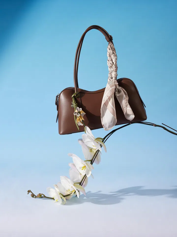 PAZZION x Jude Chan 联名 Lycorisse Orchid Leather Shoulder Bag 产品照，以浅蓝色背景搭配白色兰花营造出优雅梦幻氛围。深棕色肩背包采用简约流线轮廓设计，并在包包手柄绑上印花丝巾，搭配兰花吊饰细节，为整体造型增添浪漫艺术感。整体风格结合时尚感与柔和气质，非常适合作为母亲节送给妈妈的优雅包包礼物。