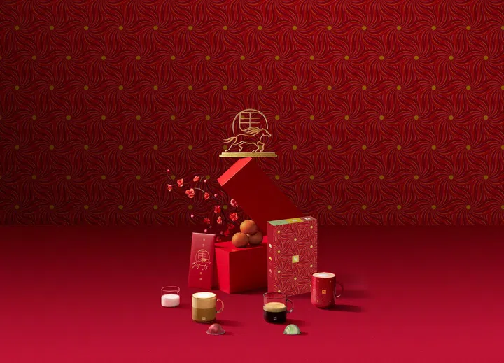 Nespresso农历新年限定系列展示图。画面以浓郁红色为主调背景，搭配金色马年装饰图腾与新春图案设计。中央陈列新年限定咖啡礼盒与节庆包装杯具，前方摆放多杯不同口味的Nespresso咖啡与胶囊，整体布置简约优雅又富有节日氛围，呈现咖啡礼盒作为新年送礼的精致选择。2026马年新年送礼推荐：拜年礼盒与新年礼物精选