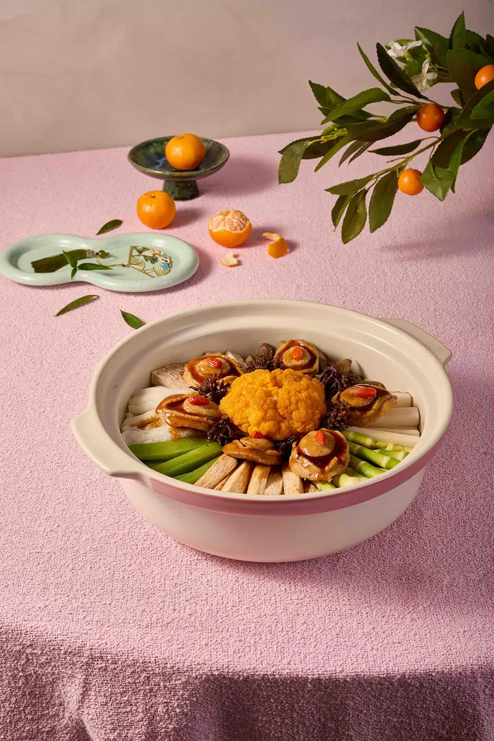 粉色桌布上摆放一盆素食盆菜，白色砂锅中以白萝卜、芦笋与多种蔬菜围成圆形摆盘，中间点缀金黄色素食配料与多块素鲍鱼，整体呈放射状排列，背景可见橘子与带叶枝条，营造农历新年节庆氛围，展现清爽风格的全素盆菜摆设。Ling Zhi 灵芝素食馆| 新加坡盆菜推荐2026 农历新年必吃餐厅与价格整理