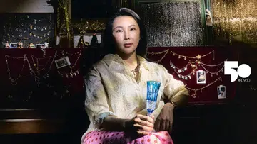 Echo Vintage Jewellery创办人Echo Wang的横幅肖像。她留着齐肩黑发，身穿米白色暗纹衬衫与粉红色红圆点半裙，佩戴项链及腕表，手持蓝色切割纹高脚玻璃杯，神情平静地望向镜头。身后的酒红色绒面展示板悬挂着多条金色、银色链饰及吊坠，穿插摆放小幅照片，上方的复古画框内密集陈列着不同款式的链饰。光线照亮她的脸庞、衬衫与蓝色玻璃杯，与深色背景形成鲜明对比；画面右侧印有白色《女友》NÜYOU 50周年标志。