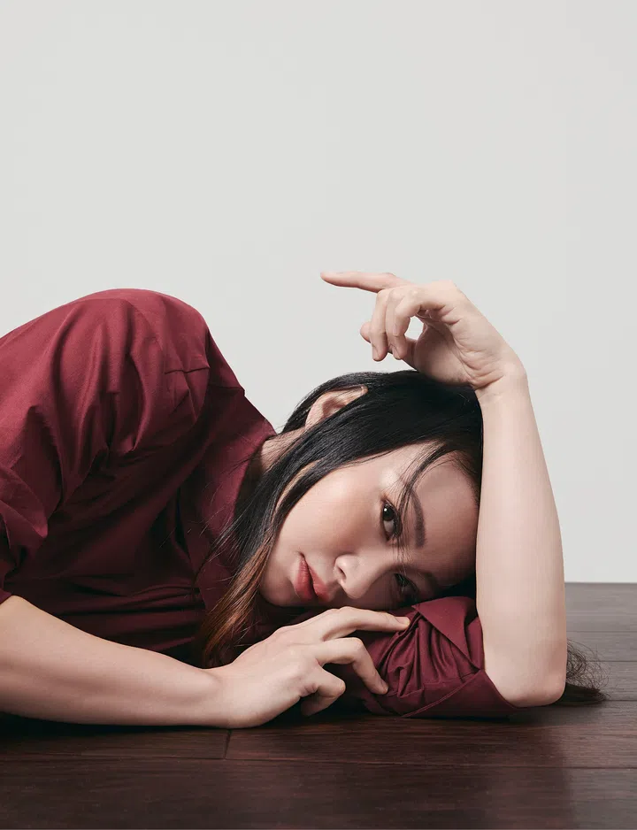 一名长发女子侧躺在深色木地板上，身穿酒红色高领长袖上衣，以手臂作为枕头，头轻靠在手臂上，直视镜头，神情平静而若有所思。另一只手自然抬起悬停于头顶上方，手指微微弯曲，营造轻盈且富有艺术感的姿态。柔和的自然光从左上方洒落，在浅灰色背景墙上形成淡淡光影，为画面增添温暖与宁静氛围。整体色调以酒红色、木质棕色及浅灰色为主，简约干净，呈现现代时尚肖像风格，同时带出放松、疗愈与沉静思考的情绪。