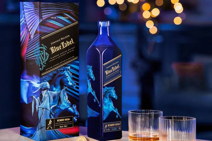 Johnnie Walker Blue Label 2026马年限定版威士忌与礼盒展示图。画面中深蓝色酒瓶与联名设计礼盒并排陈列，包装由设计师Robert Wun操刀，饰有骏马图腾与艺术感十足的蓝紫色调图案。酒瓶旁放置两只装有威士忌的水晶杯，背景为温暖的节庆灯光虚化效果，整体氛围高端优雅，呈现适合新年送礼的奢华质感。2026马年新年送礼推荐：拜年礼盒与新年礼物精选