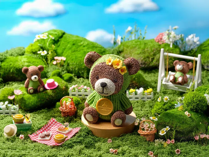 草地野餐场景中摆放一款熊造型榴莲蛋糕，穿着绿色毛衣、戴花朵头饰，周围布置小花、野餐篮与玩偶，整体氛围可爱又充满童趣。2026母亲节蛋糕推荐：精致又好吃的限定款