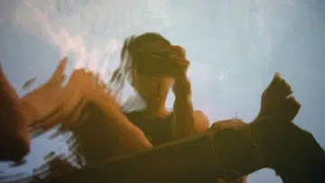 一张带有抽象感的摄影作品，画面中可见一名人物的模糊倒影，像是透过水面或反光材质拍摄而成，人物轮廓被拉伸与扭曲，形成流动感。背景呈现浅蓝色天空与柔和云影，前景带有偏暖的橙褐色调，与冷色天空形成对比。人物似乎正抬手靠近脸部，整体画面具有轻微晃动与重影效果，营造出梦幻、安静且富有情绪张力的氛围，强调光线与反射带来的视觉变化。林道锦《谁在缝隙里种光》