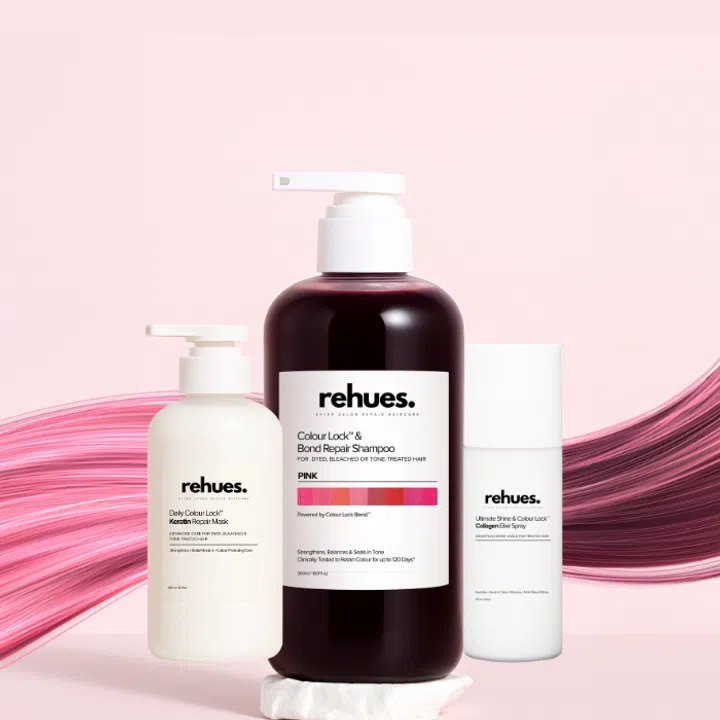 $20以下也能送出好礼：同事交换圣诞礼物精选。图中为 Rehues 染后护理系列，包括锁色修护洗发精、角蛋白修护发膜与锁色亮彩喷雾，瓶身以极简白色与深酒红配色呈现，背景搭配粉色发丝纹理，凸显染发后维持色泽与修护受损发质的专业护发主题。
