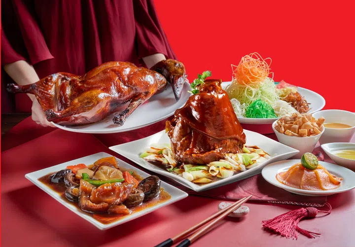 农历新年外带团圆套餐展示图，红色背景前摆放多道节庆佳肴，包括一整只酱香烤鸭由服务人员端上桌、一道浓酱焖猪手配葱段的主菜、鲍鱼香菇炖菜、橙色年糕甜点，以及一盘色彩缤纷的捞起鱼生，旁边配有脆饼与酱料碟，桌面点缀筷子与新年流苏装饰，整体呈现适合家庭聚餐的新加坡农历新年外卖团圆饭场景与热闹年味氛围。