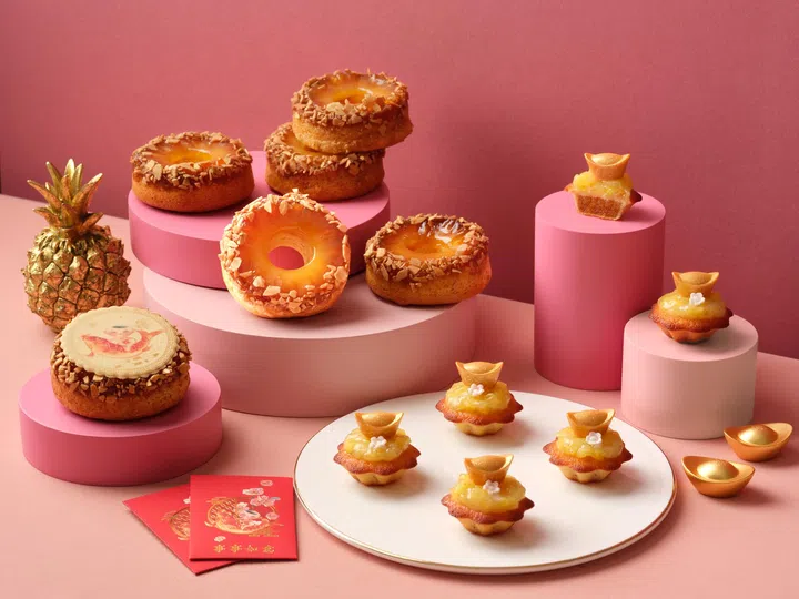 画面展示 Goodwood Park Hotel 新春主题凤梨甜点陈列在粉色背景与多层圆形展示台上，整体色调以粉红与金色为主，营造精致喜庆的新年氛围。中央展示多款圆环造型的凤梨翻转蛋糕（Pineapple Turnover Ring），表面点缀坚果碎与金黄色凤梨内馅，层层叠放在不同高度的粉色圆台上，突显甜点的立体造型与质感。右侧与前景白色圆盘上摆放多颗元宝造型的凤梨费南雪（Gold Ingots of Prosperity Pineapple Financiers），顶部装饰金色元宝与凤梨馅，寓意招财进宝。左侧可见金色凤梨装饰摆件，以及印有新年图案的圆形巧克力装饰甜点，前方摆放红色红包封套与金色元宝摆饰，整体呈现融合传统新春寓意与精致西式甜点设计的节庆甜品系列，适合新年聚会与送礼选择。2026马年年饼推荐：新春送礼与拜年点心精选