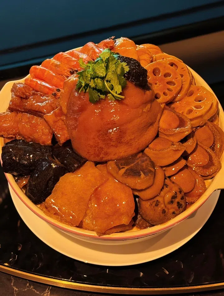 一锅满满堆叠的传统盆菜摆放在白色砂锅中，中央是一整块炆煮入味的猪肘，周围环绕着大虾、鲍鱼、海参、花胶、莲藕片与多款卤制配料，表面淋上浓郁酱汁并以香菜点缀，整体色泽红润油亮，呈现丰盛团圆的新春年菜氛围，新加坡盆菜推荐2026 农历新年必吃餐厅与价格整理