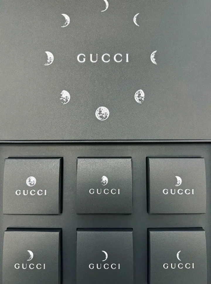 Gucci中秋礼盒采用统一的灰绿色调，打开的盒盖中央印有银白色GUCCI字样，周围以环形排列的新月、半月及满月图案装饰。下层六枚方形小盒分为两排，每盒上方印有不同形态的月亮，下方配以品牌名称。月面带有细碎纹理，整体布局整齐简洁。