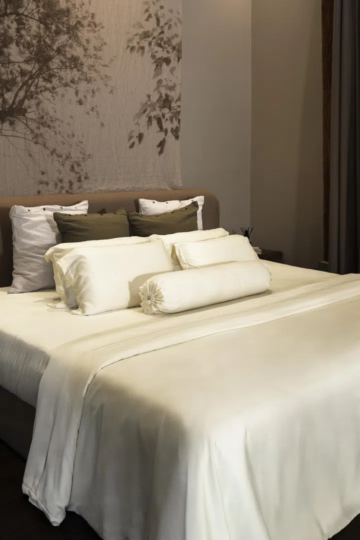 Heveya Bamboo Lyocell 床单产品照，以米白色竹纤维床单铺设整张大床，搭配同色系枕头与抱枕，整体空间以柔和暖色调营造出安静疗愈的睡眠氛围。床头背景采用树影图案设计，搭配自然感家具与低调灯光，展现Heveya主打的透气、舒适与放松睡眠体验，非常适合作为提升生活品质与睡眠幸福感的母亲节礼物。母亲节礼物推荐2026：从实用家居到时尚科技，这些礼物妈妈真的超爱！