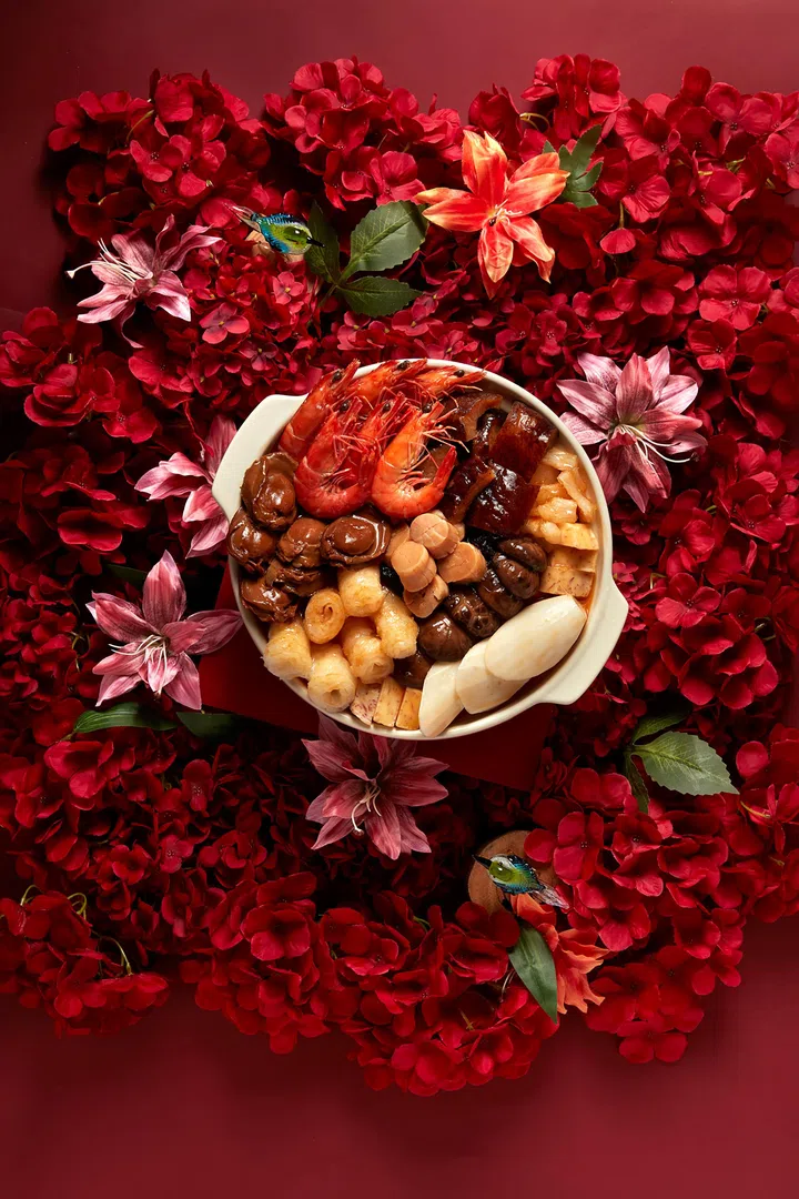 红色花卉背景中央摆放一盆潮州风味盆菜，白色砂锅中铺满整虾、鲍鱼、花胶、鱼鳔、萝卜与多款海味与配料，食材层层叠放、色泽浓亮，周围以红花与装饰营造浓厚新春节庆氛围，呈现适合农历新年团圆聚餐的盆菜摆设，新加坡盆菜推荐2026 农历新年必吃餐厅与价格整理。