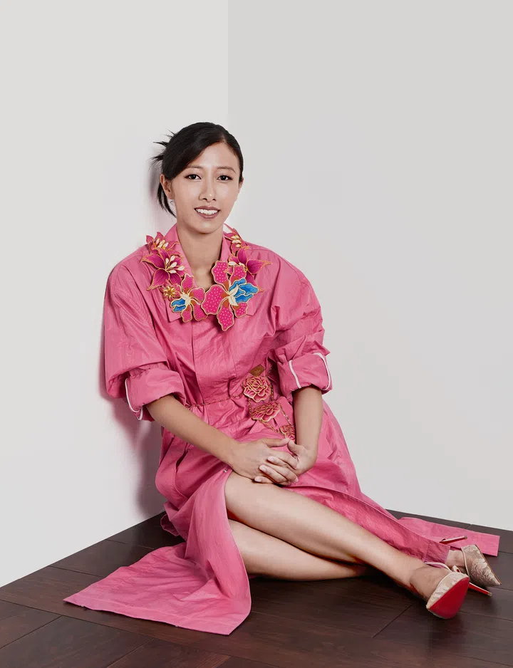Serena Wong 穿着粉色长裙坐在地板上，颈间佩戴由多枚花卉胸针组合而成的项链，腰间点缀着三件套胸针（kerongsang），作品皆为她亲手创作。