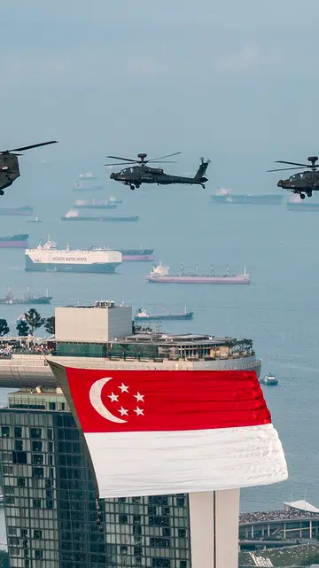 新加坡滨海湾（Marina Bay）上空，三架新加坡空军军用直升机编队飞行，其中一架CH-47支奴干直升机悬挂着一面巨大的新加坡国旗，旗帜迎风展开，鲜红与白色在空中格外醒目。前景可见滨海湾金沙酒店（Marina Bay Sands）标志性的三座塔楼，国旗正从其中一座塔楼前方掠过。远处的新加坡港口布满数十艘货轮与集装箱船，平静的海面延伸至地平线，天空带有淡淡薄雾，营造出庄严而壮观的氛围。整幅画面展现新加坡国庆庆典彩排期间的空中飞行表演，同时凸显这座海港城市的天际线与繁忙港口景观。