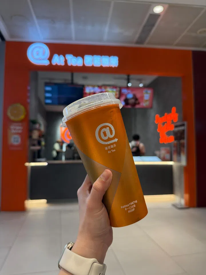 新加坡 At Tea 署茗职茶门店前，一只手举着一杯橙色透明杯装茶饮，杯身印有品牌标志，背景为橙色主题的店铺招牌与柜台，灯光明亮，营造年轻时尚的手摇饮氛围，展现明星萧敬腾创立的茶饮品牌。