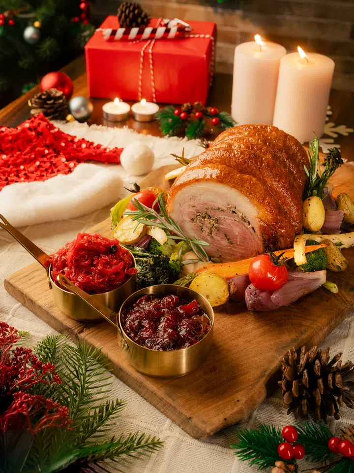 金黄酥脆的圣诞烤猪肉卷摆放在木板上，搭配烤蔬菜、蔓越莓酱与腌制红甘蓝，背景有圣诞礼物、松果、蜡烛与节日装饰，呈现 Surrey Hills Grocer 的圣诞外带料理。2025圣诞外带套餐推荐：在家享用超值又美味的圣诞大餐